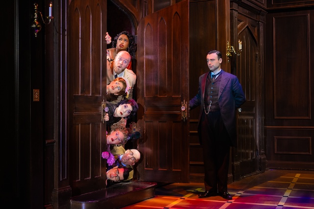 Seven actors lay stacked on top of each other horizontally, peeking out from behind an open door. Only their
surprised
faces are visible outside of the door frame. Across from them, a man in a suit has one arm outstretched, having
just opened the door